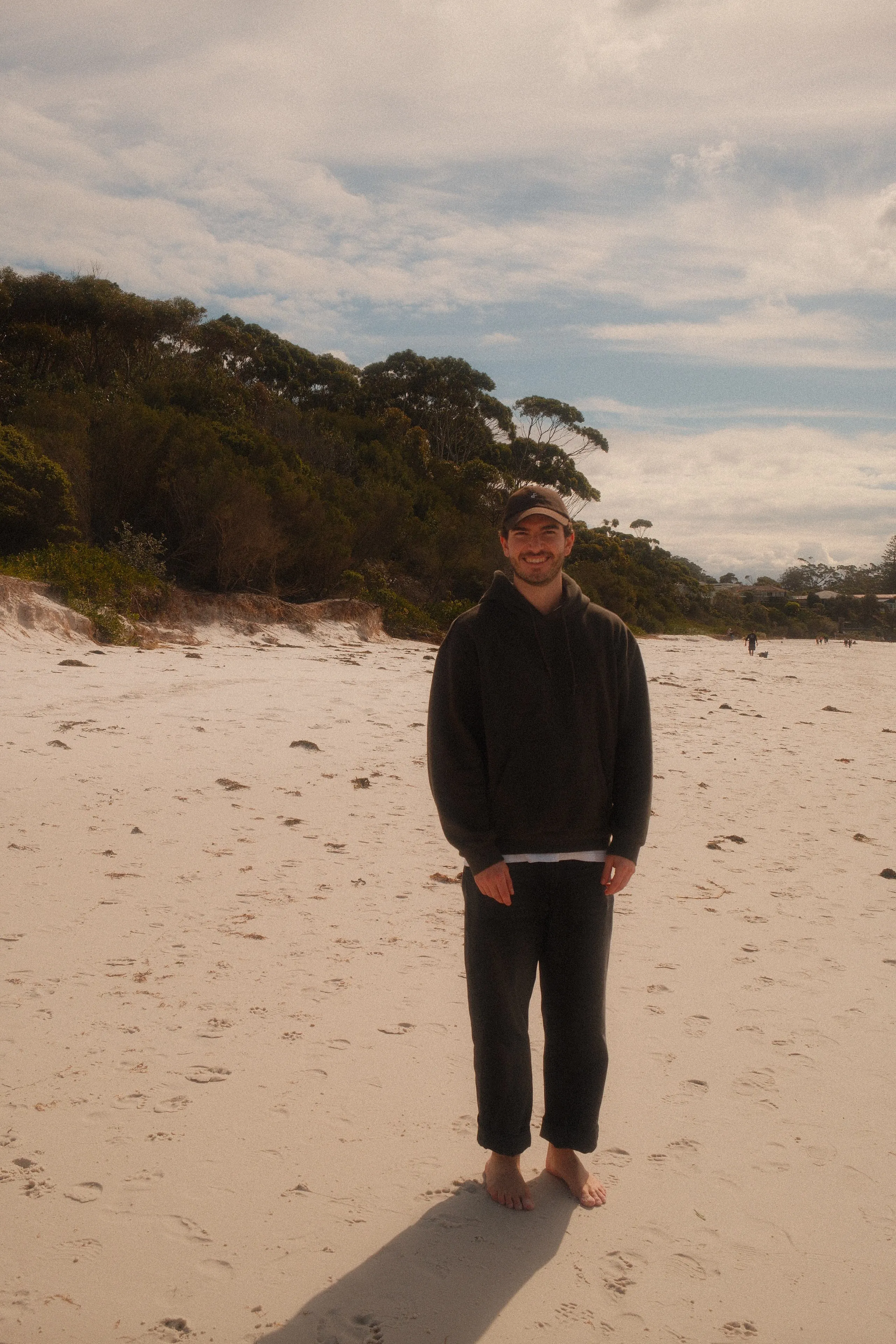 Damian standing barefoot on a wide sandy beach in a dark hoodie, smiling