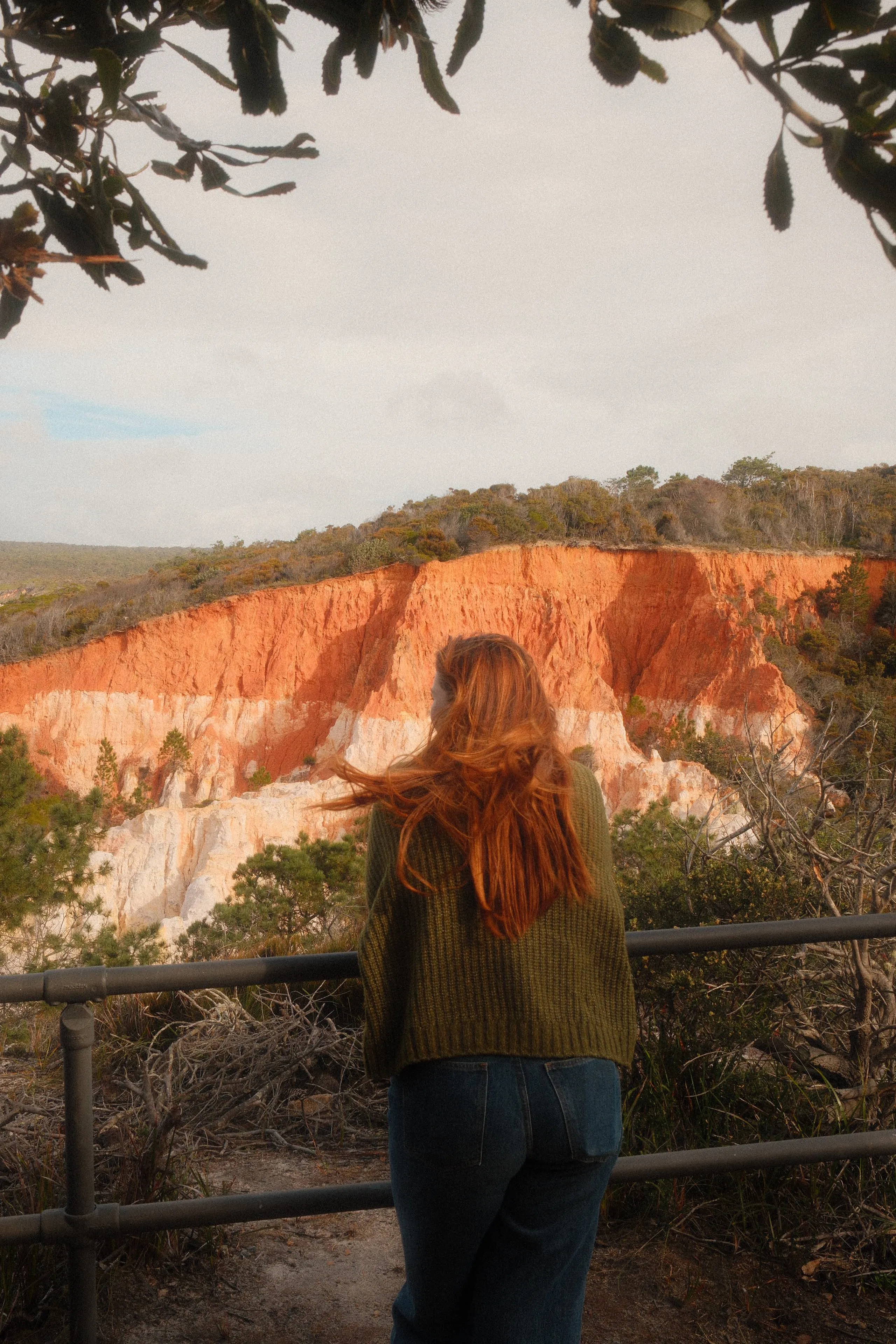 A woman with long red hair blowing in the wind, looking out at orange and white eroded cliffs