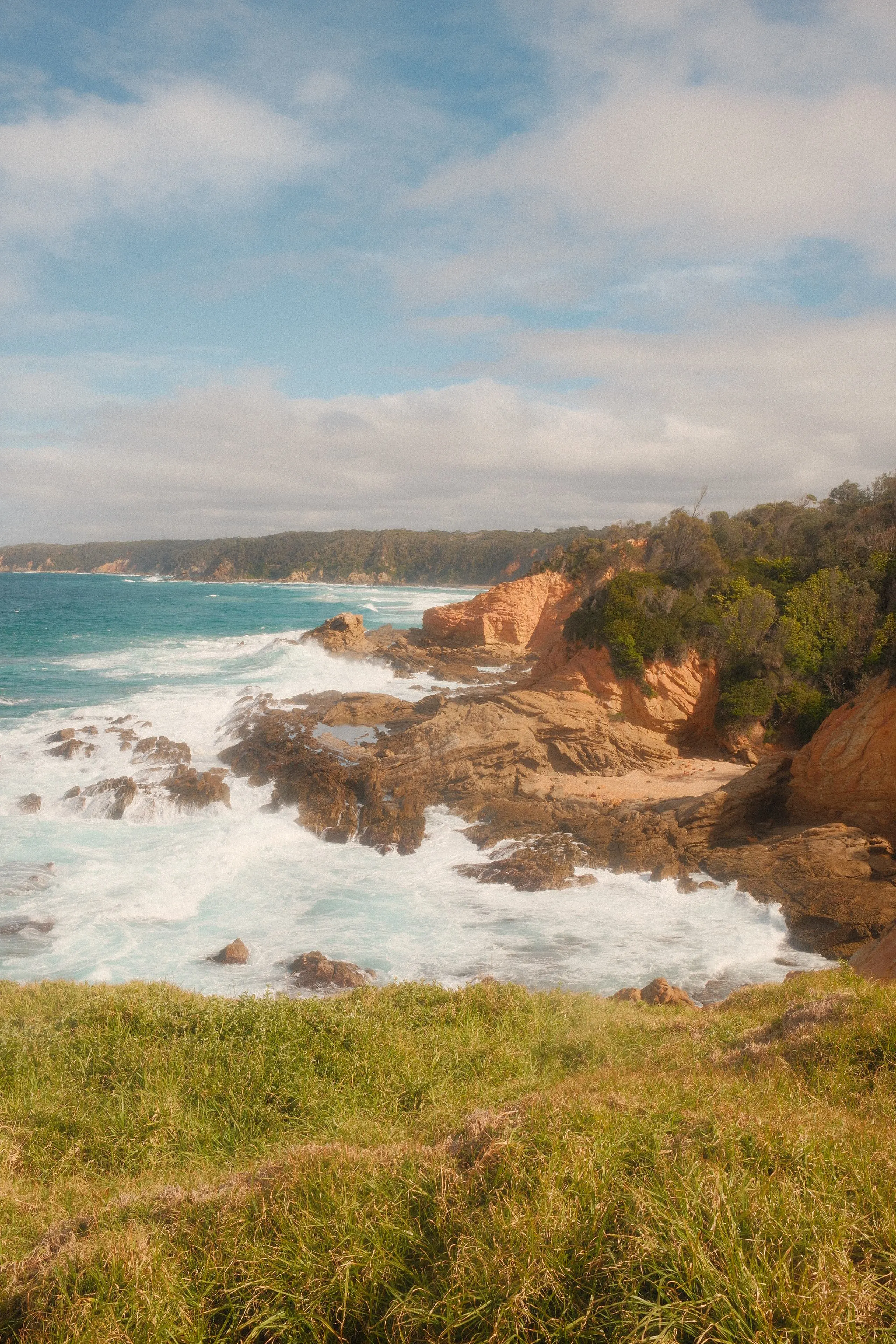 Waves breaking white over orange rocks below a grassy headland