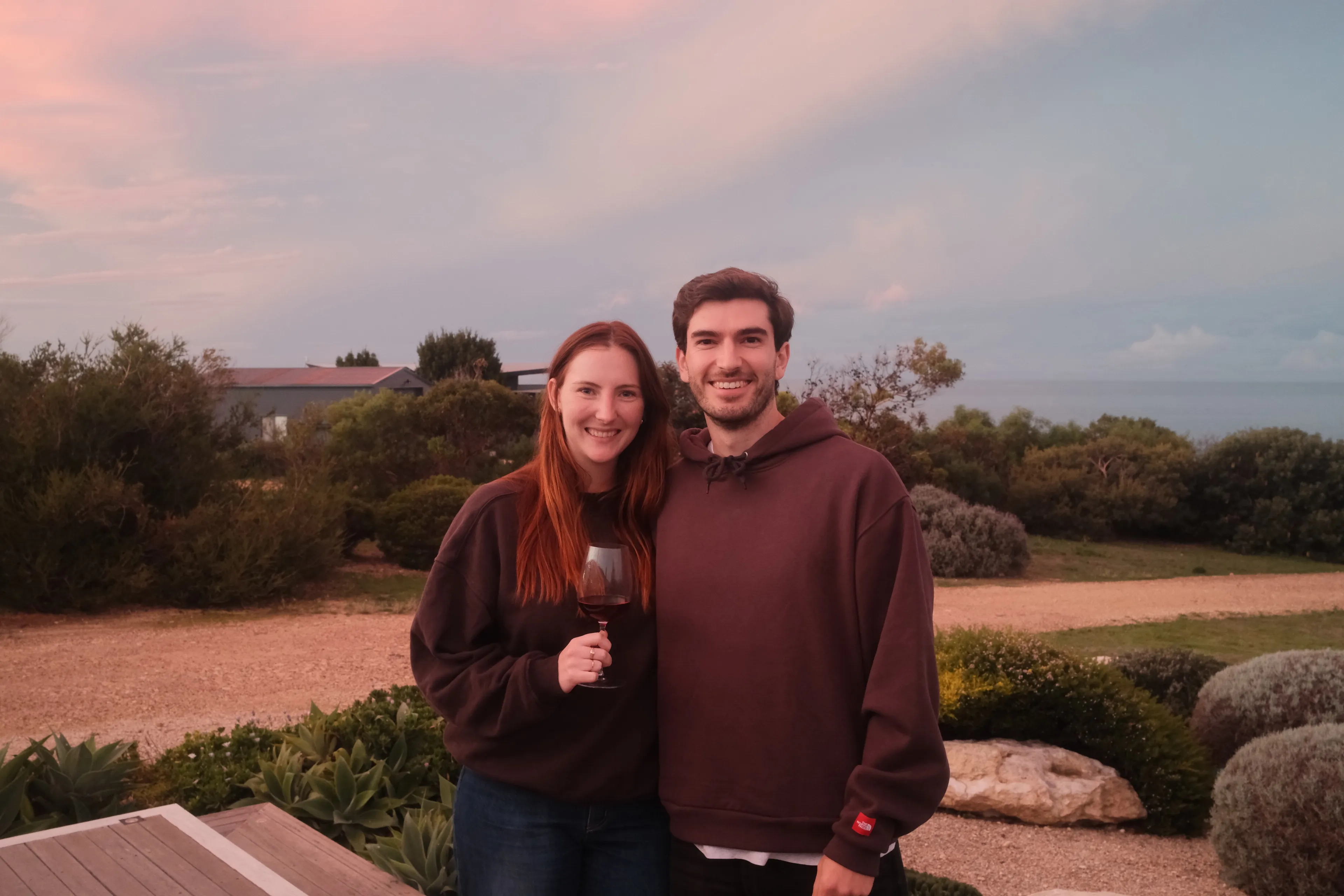 Damian and a woman holding a glass of red wine, smiling in a garden under a pink dusk sky