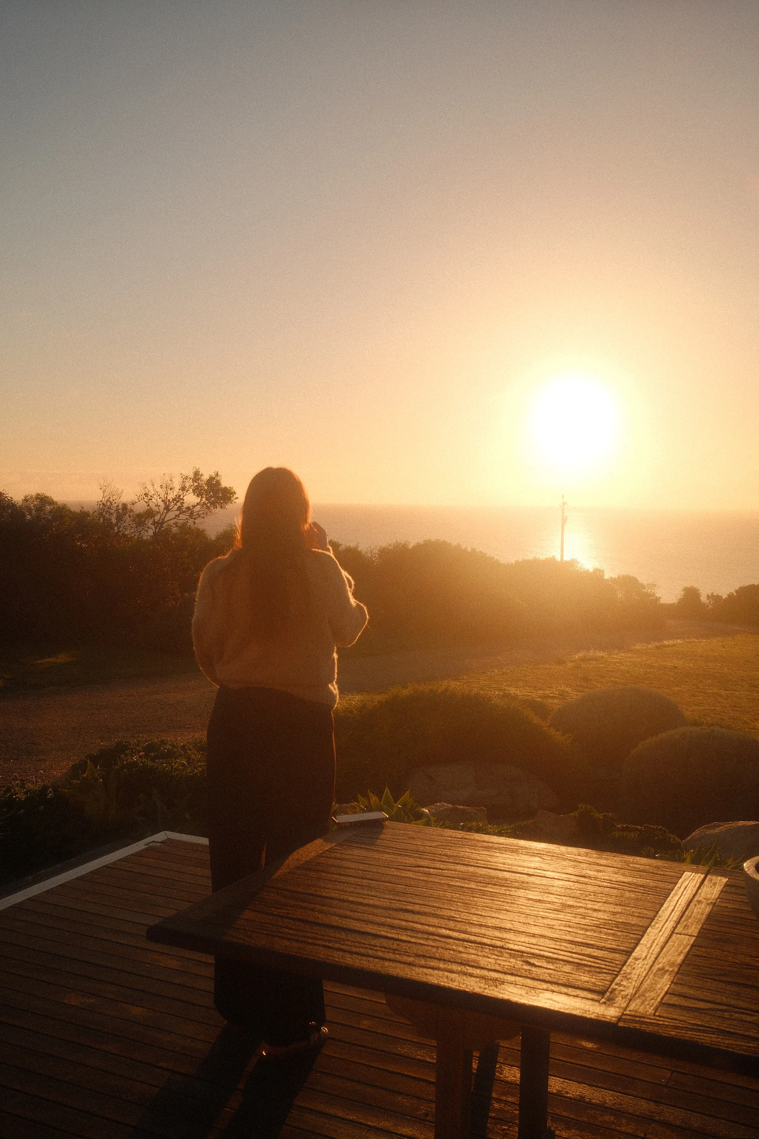A woman on a timber deck looking out at the sun setting over the ocean