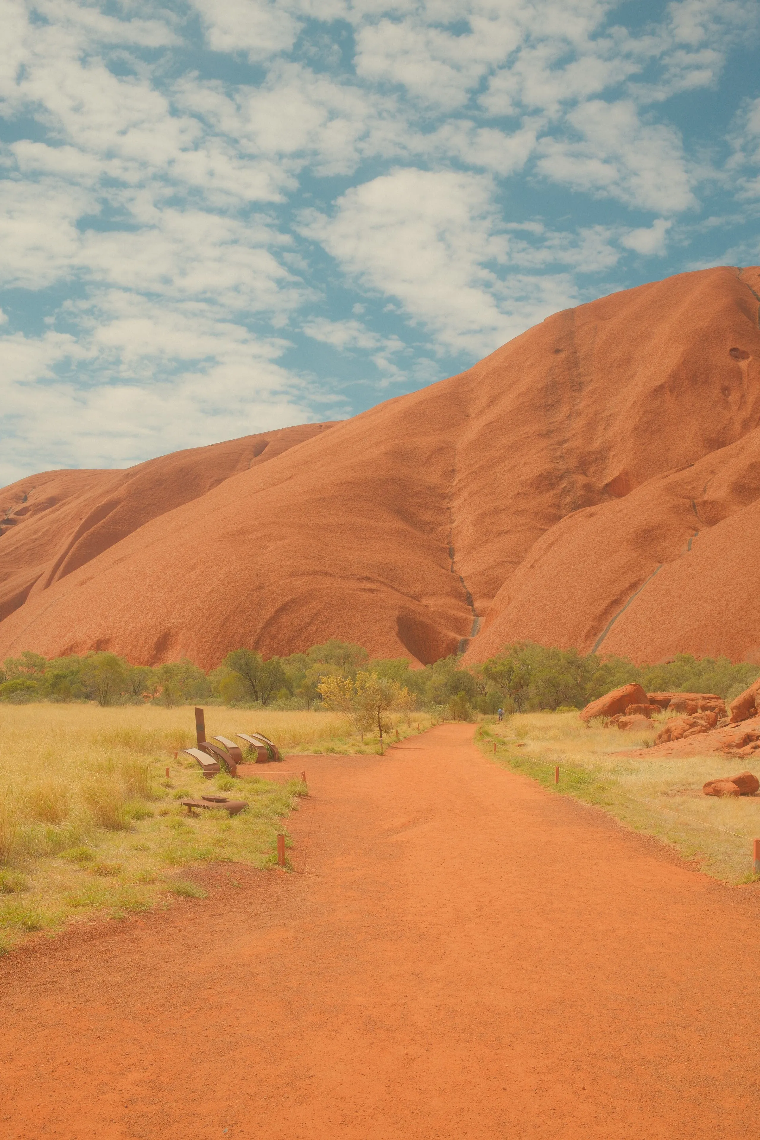 A red dirt path leading to the base of Uluru under a sky of scattered white cloud